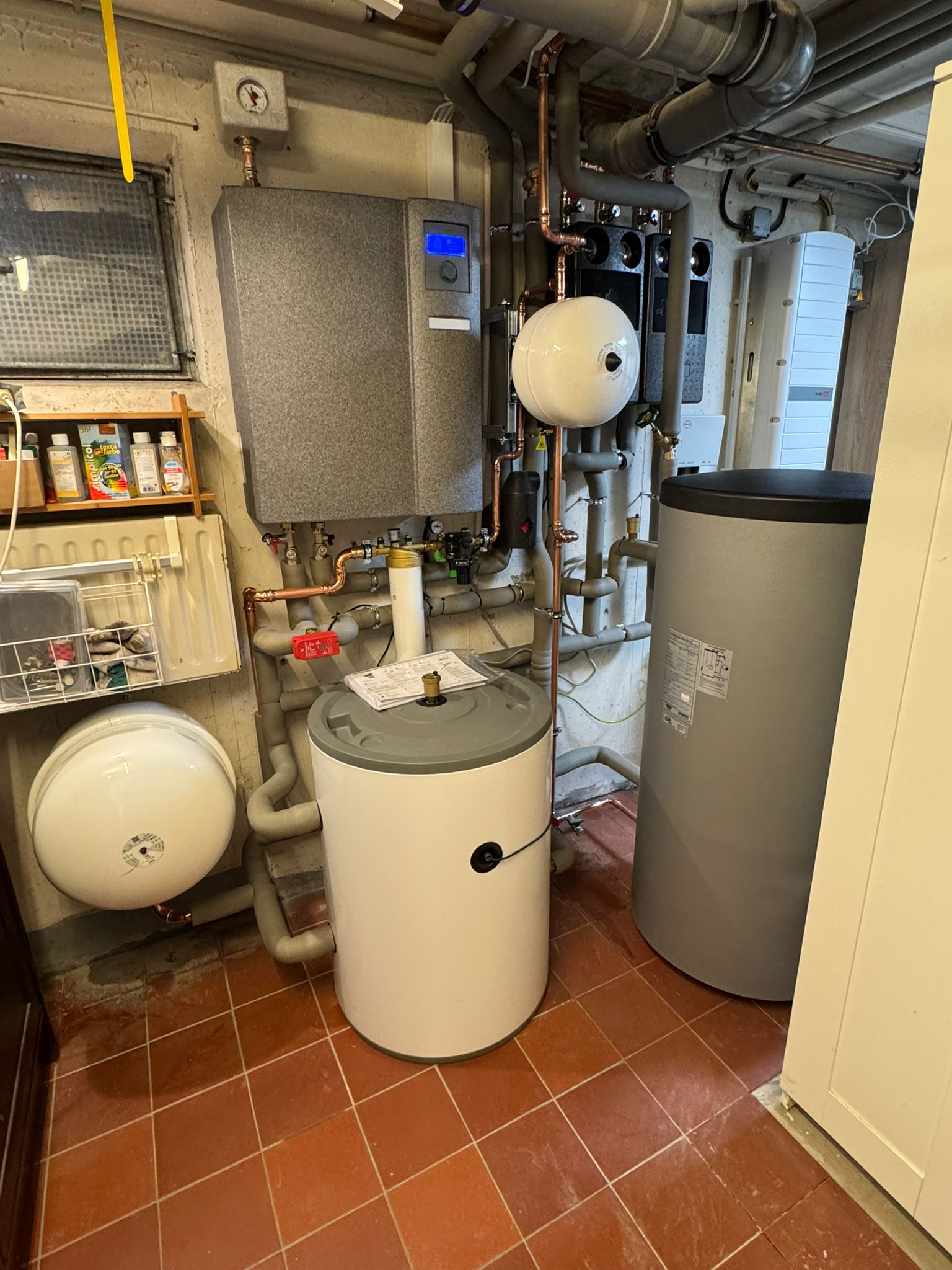 Technikraum mit Pufferspeicher, Wandgerät, zwei Ausdehnungsgefäßen und zahlreichen Rohrverbindungen.
